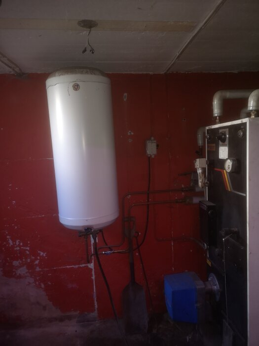 Boiler with a white cylindrical water heater tank mounted on a red-painted wall, accompanied by various pipes and equipment.