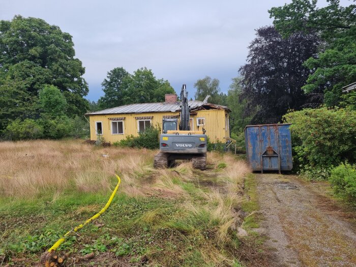 Grådassig grävmaskin framför ett gult förfallet hus med skadat tak och hög växtlighet runtom. Bredvid huset står en blå metallcontainer.