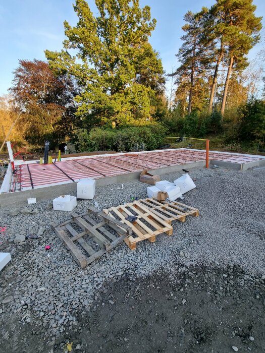 Byggarbetsplats med isoleringsmaterial, två träpallar och grund för hus under uppförande, omgiven av grönska och höga träd.