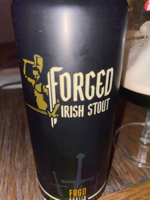 Burk med Forged Irish Stout öl på ett träbord, bredvid ett glas delvis fyllt med öl.