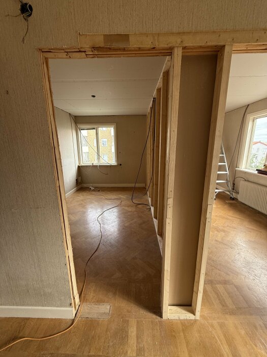 Två rum under renovering med nyligen uppsatta väggar, synliga elinstallationer och en stege i bakgrunden. Båda rummen har trägolv och fönster.