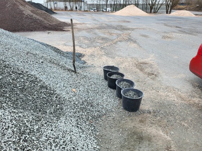 Fem hinkar fyllda med grus står på marken bredvid en stor grushög. Del av en röd bil syns till höger.