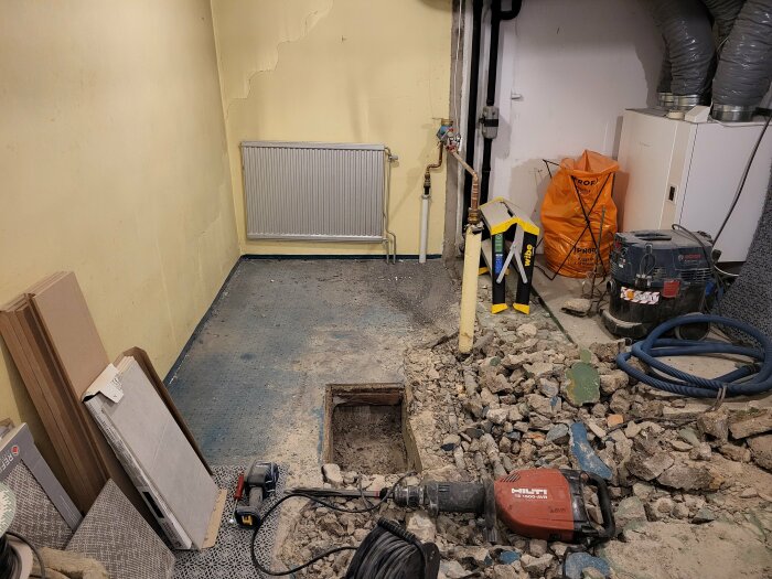 En renovering pågår i ett pannrum med borttaget golv, synliga rör, ett element, byggmaterial och verktyg inklusive en Hilti TE 1000-AVR i bildens förgrund.