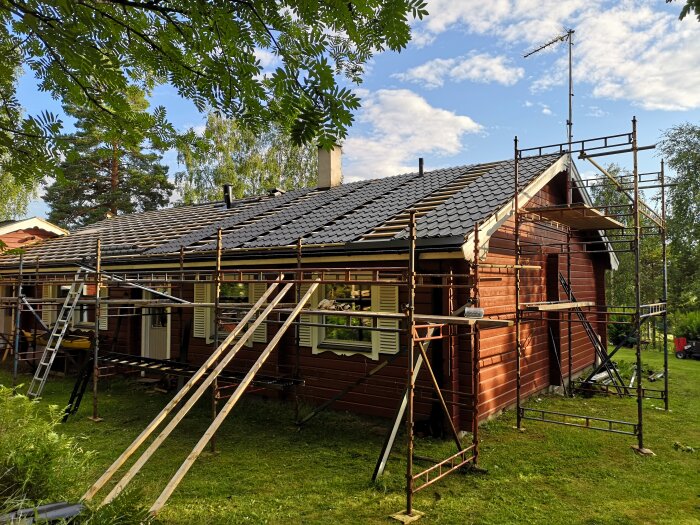 Byggnadsställningar runt ett träfölat hus, förberett för renovering och målning, med träplankor och stegar under en klar himmel.