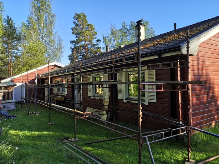 Byggställning av Haki typ framför ett rött trähus med vitt fönster, grönt gräs och träd i bakgrunden.