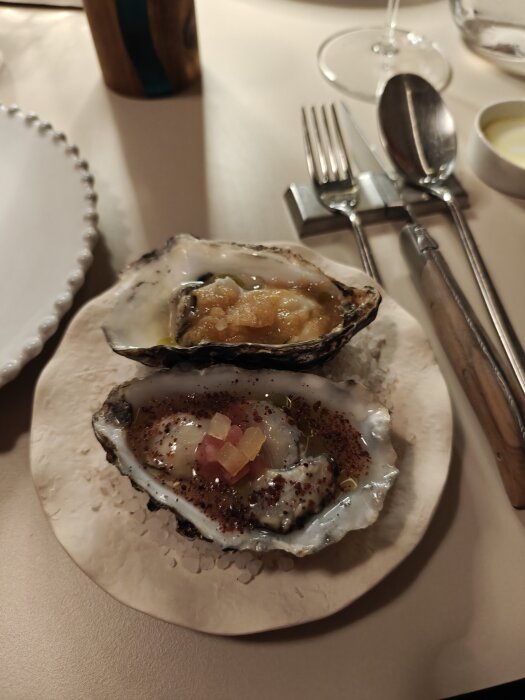 Två gratinerade ostron serverade på en fin restaurang med bestick och vinglas i bakgrunden.