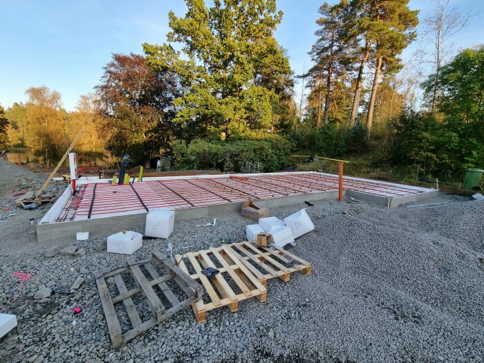 Byggarbetsplats med grundläggning för ett hus. Synliga rör för el och vatten samt ett trägolv under uppbyggnad, med träd och natur i bakgrunden.