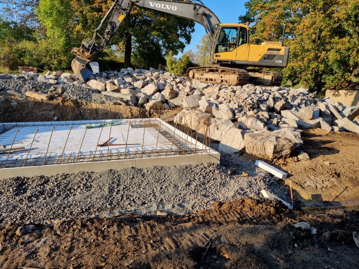 Grävmaskin står på en stor hög med stenblock bredvid en gjuten betonggrund med armeringsjärn på en byggarbetsplats omgiven av gröna träd.