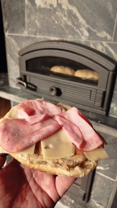 Hand håller en smörgås med skinka och ost framför en kakelugn med nybakat bröd inne i ugnen.