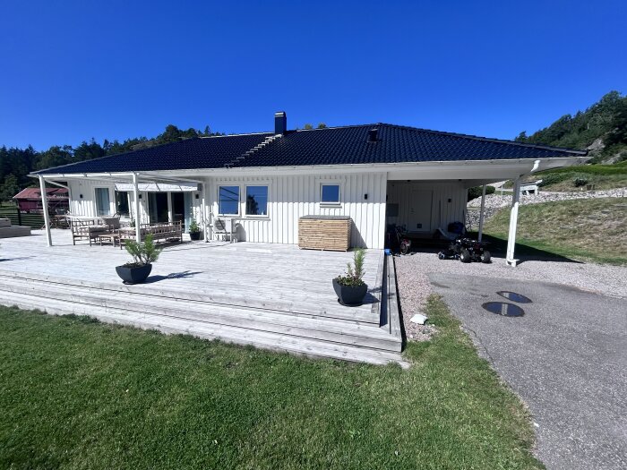 En vit villa med altan, blommor i krukor och en carport under ett svart tak. Grönska och blå himmel syns i bakgrunden.