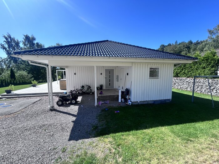 Vit carport med sadeltak gjord av mörkblå takpannor, med cyklar och leksaker under taket och grönska, träd samt ett stängsel i bakgrunden under en klarblå himmel.