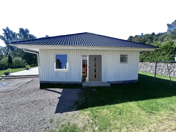 Vit carport med dörr och två fönster som ska byggas om till sovrum, sett från gårdsplan med grus och grön gräsmatta i en trädgård med stenmur.