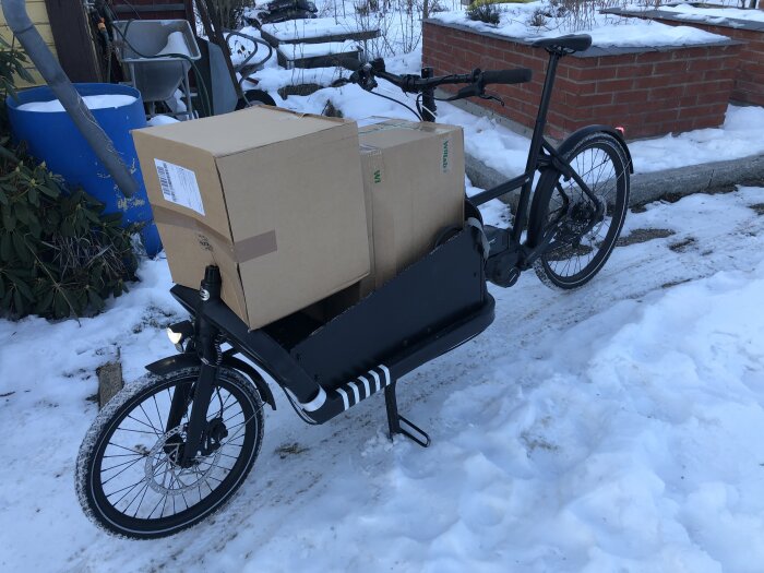 Svart lastcykel med stora kartonger framtill, parkerad på en snötäckt mark.