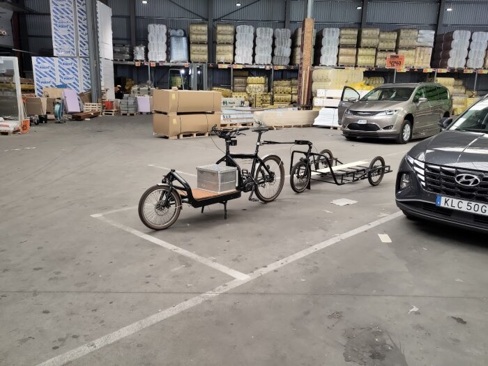 Lastcykel med släp parkerad inne i en byggvaruhandel, omgiven av lastpallar och olika material. Flera bilar är parkerade, inklusive en Hyundai och en silverfärgad van.