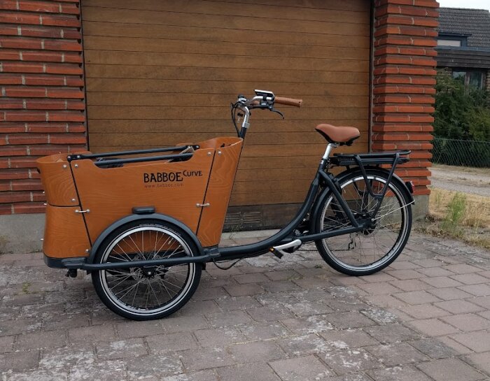 Lastcykel av märket Babboe Curve parkerad framför en garageport. Cykeln har stort lastutrymme fram och sadel samt handtag i brunt läder.