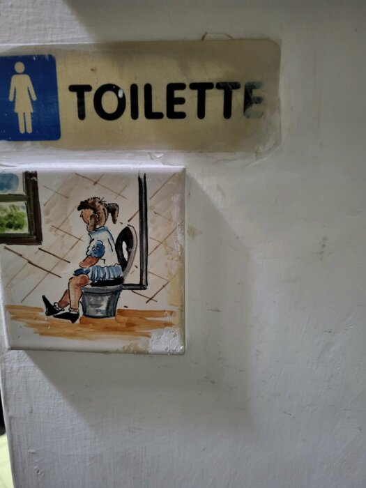 Skylt med texten "TOILETTE" och en målning av en flicka som sitter på en toalett, placerad på en vägg.