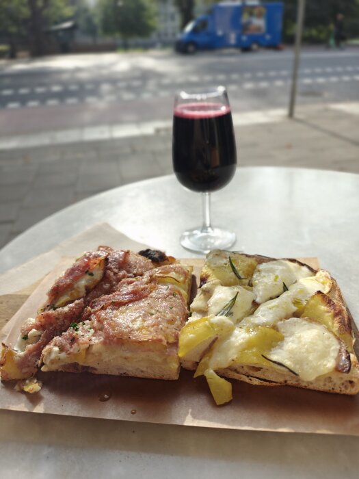 Två bitar pizza med olika toppings och ett glas rött vin står på ett bord utomhus med en gata i bakgrunden.