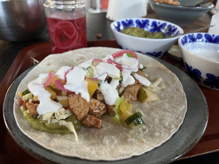 Grillad kyckling taco med picklad rödlök, vitlökssås, grönsaker, salsa, riven ost och guacamole på en mjuk tortilla, omgiven av skålar med tillbehör.