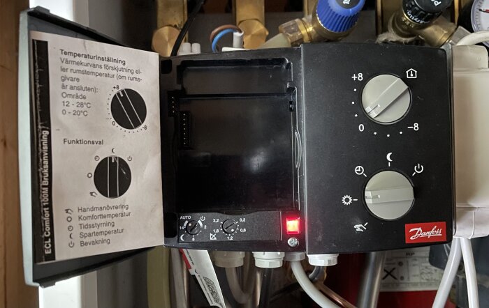 Termostat för fjärrvärme med två inställningsrattar och en LED-lampa. Instruktioner för temperaturinställning och funktionsval på insidan av luckan.