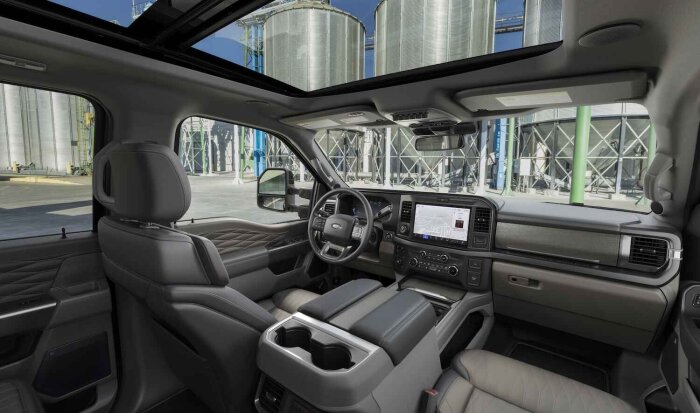 Interiör av 2025 Ford Super Duty Platinum med grå vadderade säten, central navigationsskärm och panoramasoltak, med industribakgrund utanför fönstren.