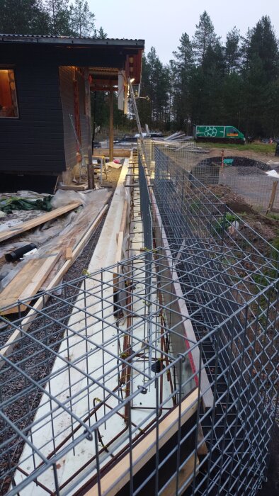 Byggprojekt med gabioner och formsättning samt byggmaterial vid ett hus, omgivet av träd.