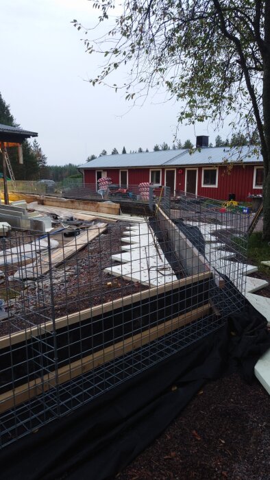 En byggarbetsplats med gabioner och träramar under konstruktion framför en röd byggnad på landsbygden.