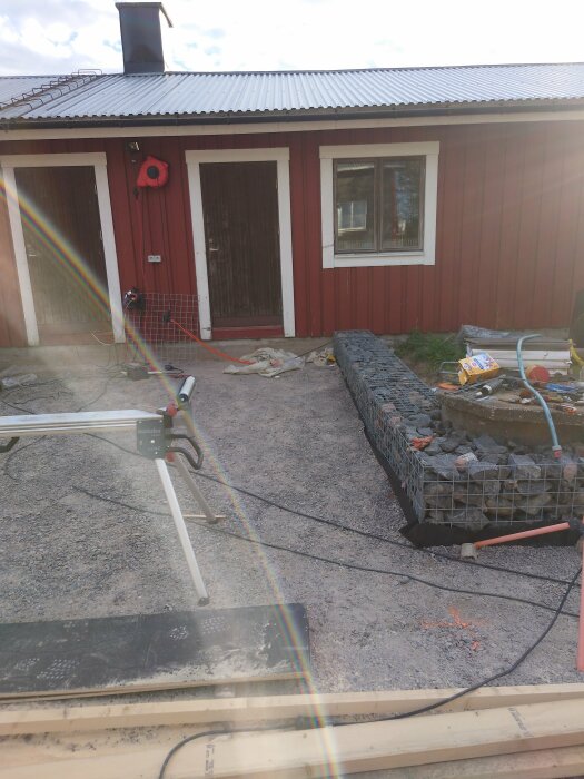 En byggarbetsplats med gabioner fyllda med stenar framför en rödmålad byggnad. Verktyg, byggmaterial och en såg på ett stativ syns också.