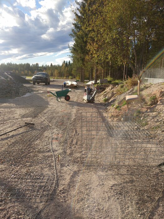 Byggarbetsplats utomhus med trådformade gabioner, verktyg, skottkärra, staplade material och en bil i bakgrunden under en klar himmel.