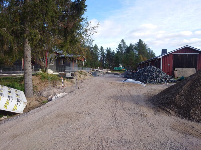 En byggarbetsplats med grusväg, hög med stenar, röda byggnader i bakgrunden samt diverse byggmaterial på marken och en gran i förgrunden.