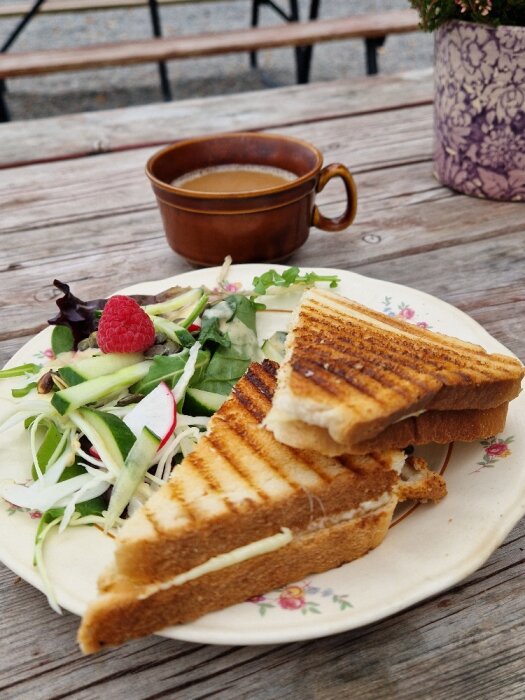 Grillad toast med ost och skinka, serverad med en sallad innehållande rädisa, pumpafrön och hallon. Kaffe i en brun kopp på ett bord utomhus.