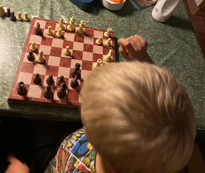 Ett barn spelar ett parti schack vid ett bord med grön duk, med schackpjäser i både vitt och brunt placerade på brädet.