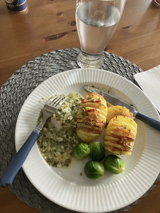 Tallrik med torskrygg i bechamelsås med spenat och keso, gratinerad med parmesan, hasselbackspotatis med bacon och brysselkål, bredvid ett glas vatten.