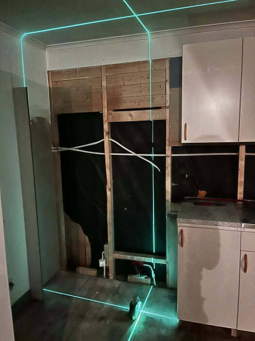 Renovering av en vägg med exponerade reglar, gröna laserlinjer projicerade på väggen och golvet, köksskåp synliga till höger.