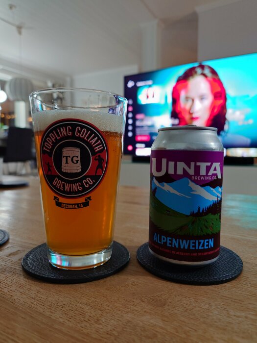 Ett glas med öl från Toppling Goliath Brewing Co och en burk Alpenweizen veteöl från Uinta Brewing Co på ett träbord med en bakgrund av en påslagen tv.