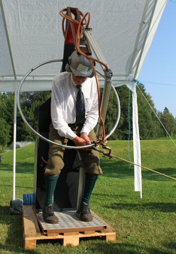 Man i traditionell klädsel står på en simulator eller maskin i en öppen gräsyta, under ett tält med kabeltråd och metallbåge runt honom.