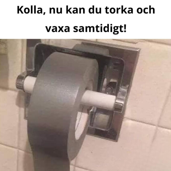 Rulle med silverfärgad tejp som är placerad i en toalettpappershållare på en kaklad vägg, med texten "Kolla, nu kan du torka och vaxa samtidigt!" ovanför.