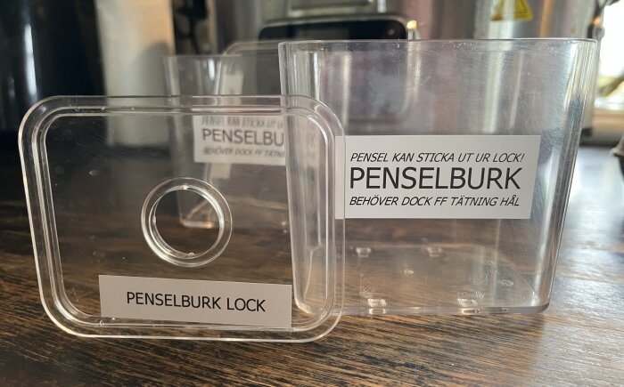 Plastpenselburk med texten "PENSELBURK" och ett lock bredvid med hål och texten "PENSELBURK LOCK". Locket har en markerad öppning för pensel.