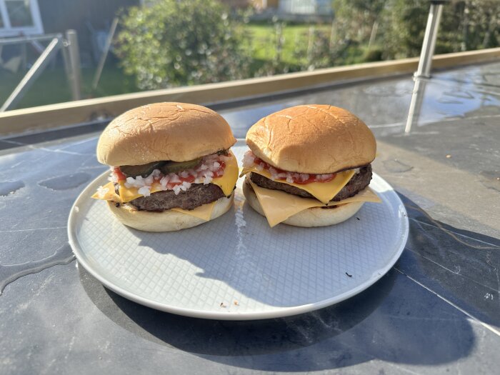 Två ostburgare med picklad gurka, lök och ketchup på en tallrik utomhus, troligtvis från en grillning.