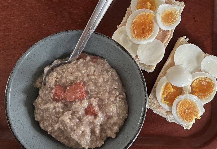 En skål havregrynsgröt med medkokta jordgubbar och två äggmackor med kaviar serverade på en mörkbrun bricka.