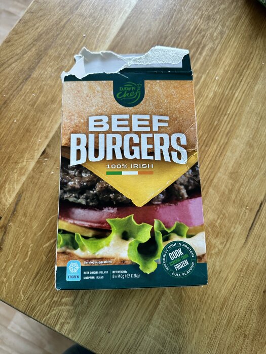 Förpackning av Dawn Chef Beef Burgers, 100% irländskt nötkött, på ett trädgårdsbord. Förpackningen är delvis öppen, innehåller 8 burgare om 140 g vardera.