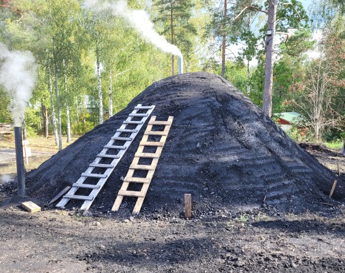 Två stegar lutar mot en stor rykande kolhög i ett skogsparti med träd och grönska i bakgrunden.