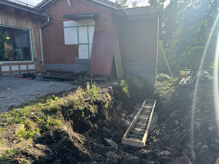 Formar för en gjuten sockel vid sidan av ett hus, omgiven av grävmaterial och vegetation. Ett rött tegelhus med ett träpanelhus syns i bakgrunden.