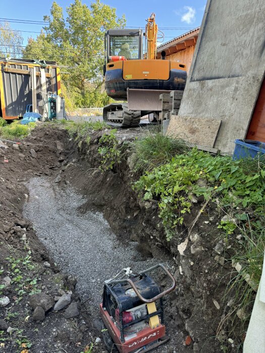 Grävmaskin och utgrävd dikesliknande sänka fylld med sten, förberedd för gjutning, med verktyg och byggmaterial runtom samt en tegelvägg i bakgrunden.