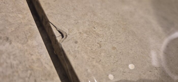 Kantstött kakelplatta i ett badrum med en synlig spricka nära brunnen, förmodligen orsakad av ett tappat duschhandtag i metall.