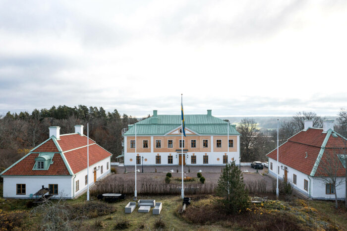 Huvudbyggnaden på Mems slott med två flygelbyggnader, grusplan, flaggstång och omgivande natur.