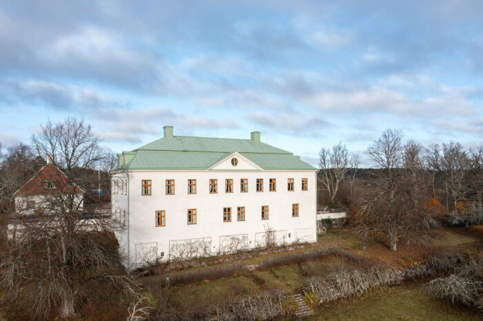 Bild på en vit herrgårdsbyggnad (Mem slott) med grönt tak och många fönster, omgiven av träd och grönska under en molnig himmel.