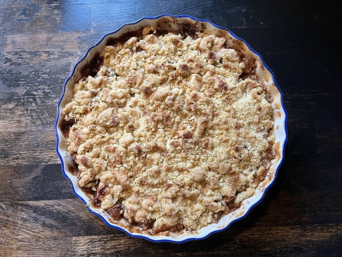 Nygräddad smulpaj med äppelbitar i en vit-randig pajform, serverad på ett mörkt träbord.