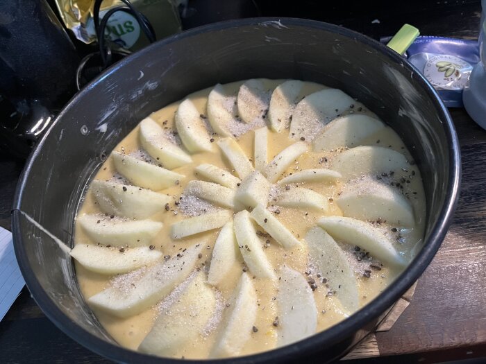 Ogräddad äppelkaka i en springform med äppelklyftor arrangerade på smeten, strödda med kardemumma och socker.