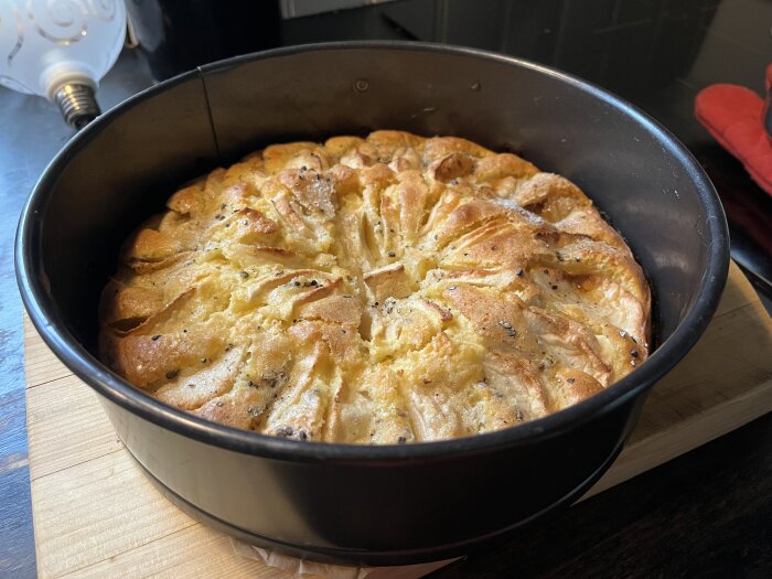 Äppelkaka med citron och kardemumma i en rund bakform, toppad med äppelklyftor fint placerade och beströdd med kardemumma och sockerblandning.