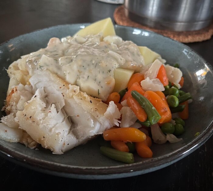 Tallrik med stekt torsk, kokta potatisar, kokta grönsaker (ärtor, morötter, gröna bönor), och äggsås med dill.